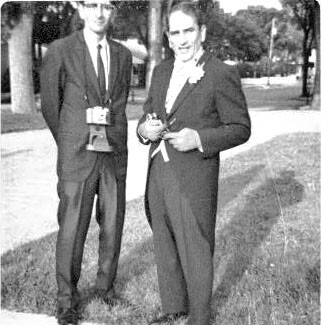 Two suited professionals standing on grass outdoors; man left wears a badge, man right holds a rolled document.