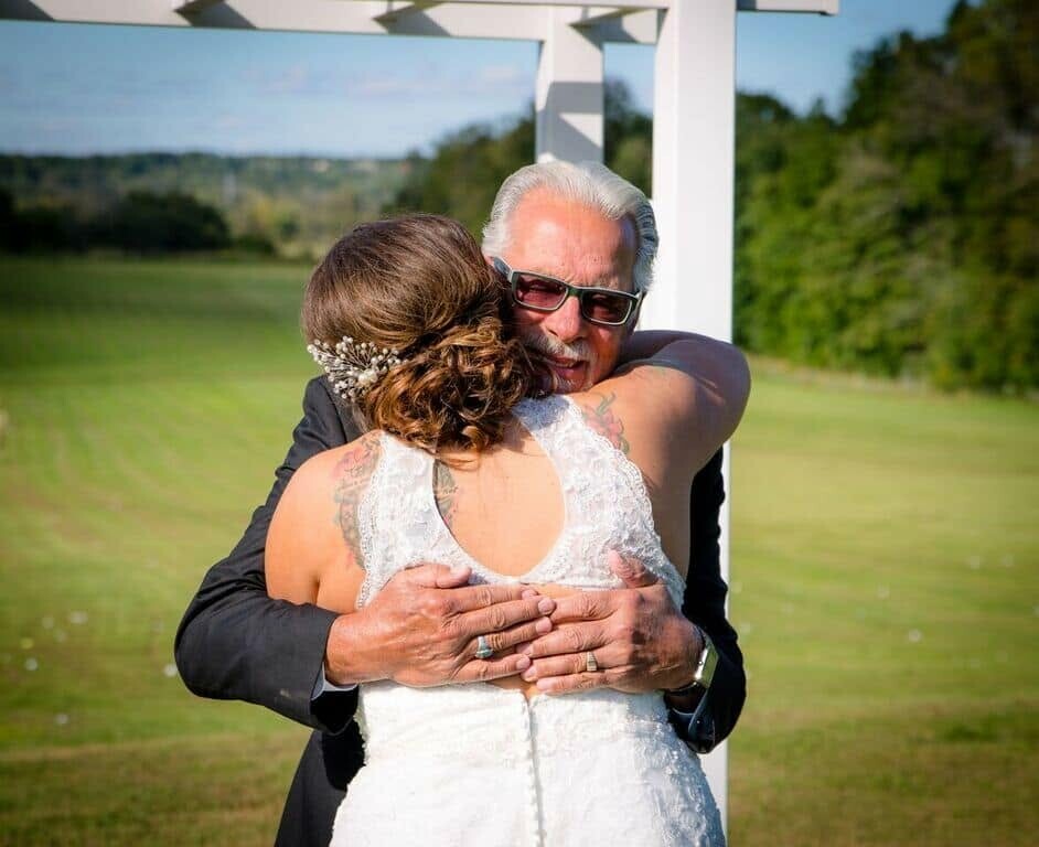 Bride in a lace wedding dress embraces an older man in a dark suit outdoors by a white arch, both smiling and holding hands over her back.