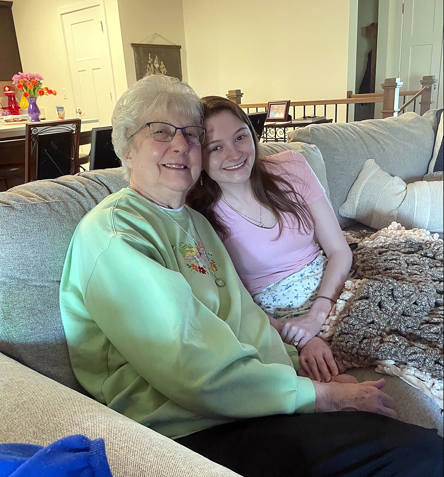 Smiling grandmother and granddaughter sit closely on a gray couch in a cozy living room, posing for a selfie.