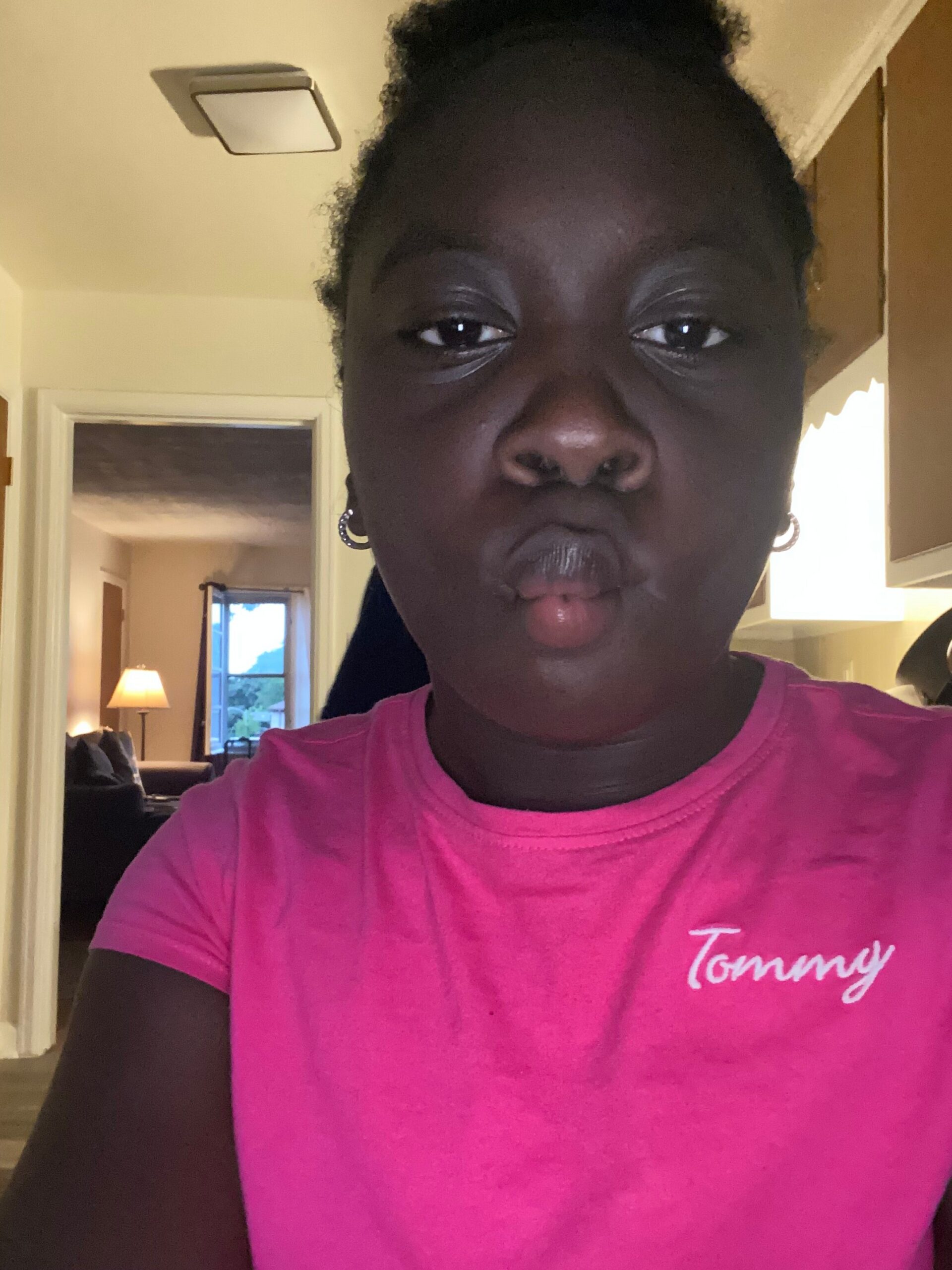 Close-up selfie of a person with dark skin, pursed lips, wearing a bright pink shirt with white cursive text, in a home interior.