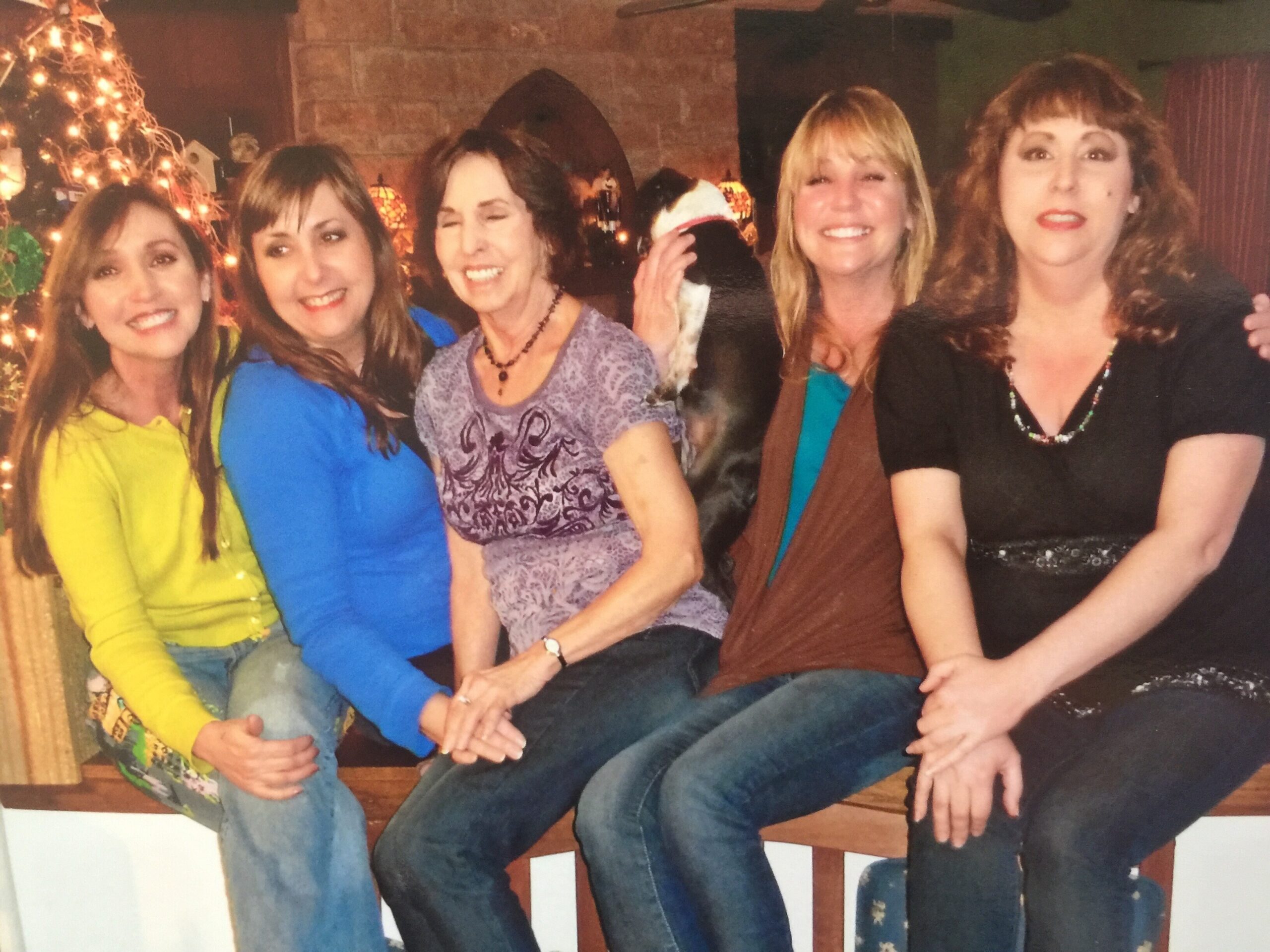 Five women sit close together on a bench indoors, smiling at the camera with holiday lights in the background.