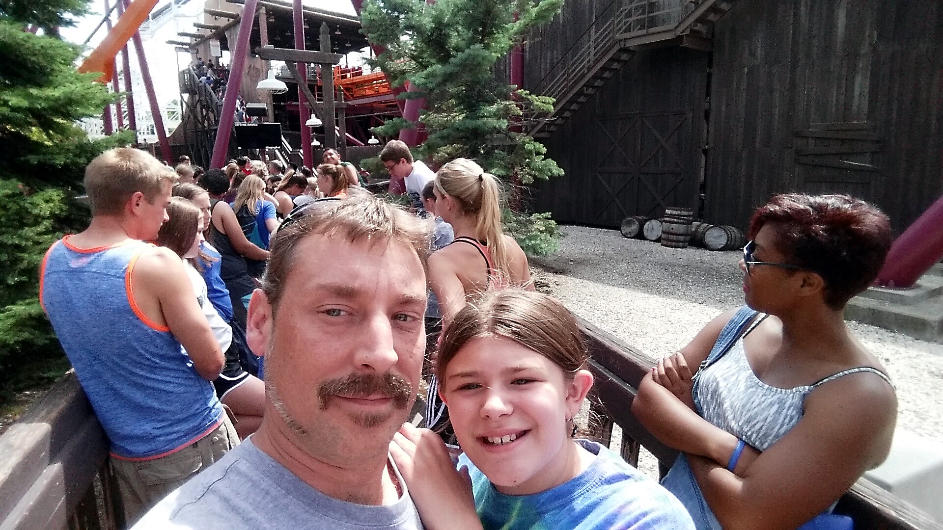 Man and girl in foreground taking a selfie as they stand in a long queue for a wooden roller coaster ride