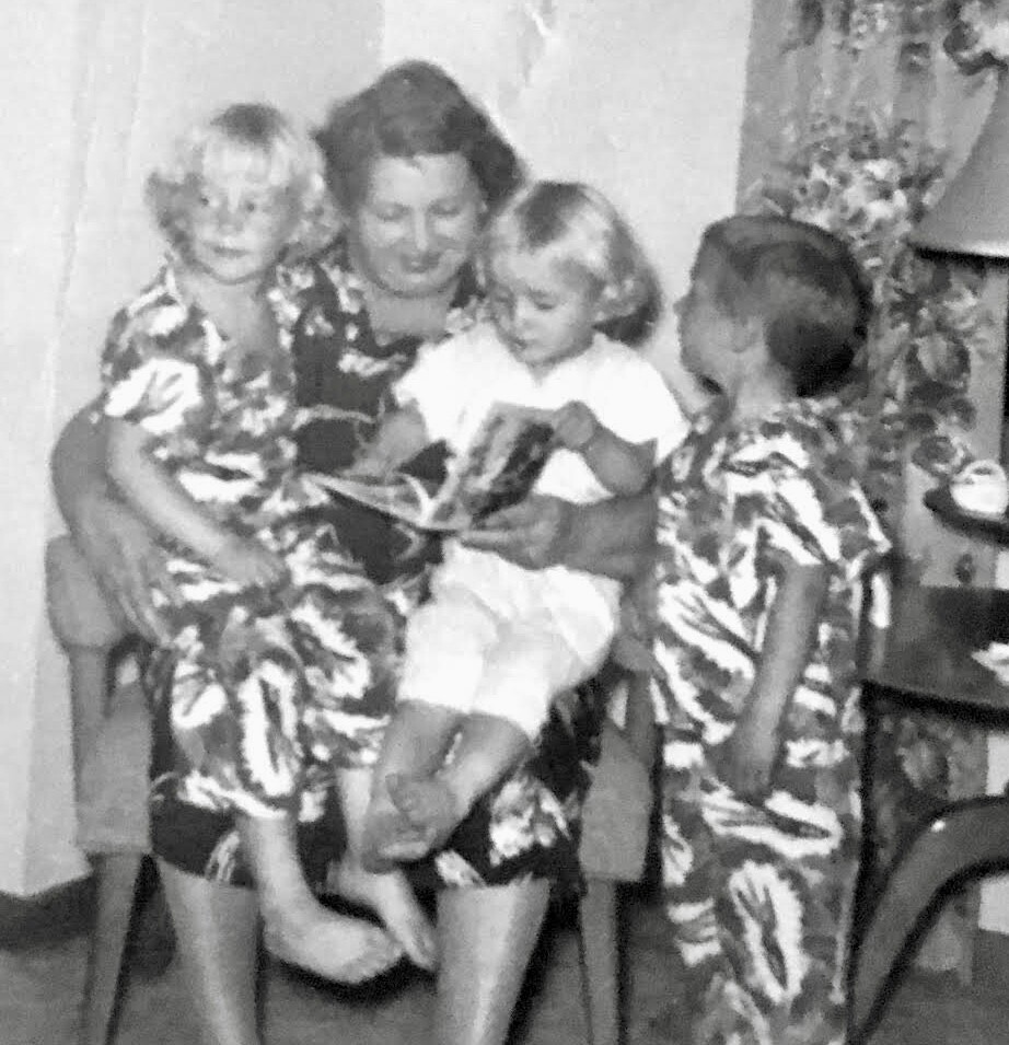 A woman seated with three children indoors, reading a book as they gather around her; one child on her lap and two standing nearby.