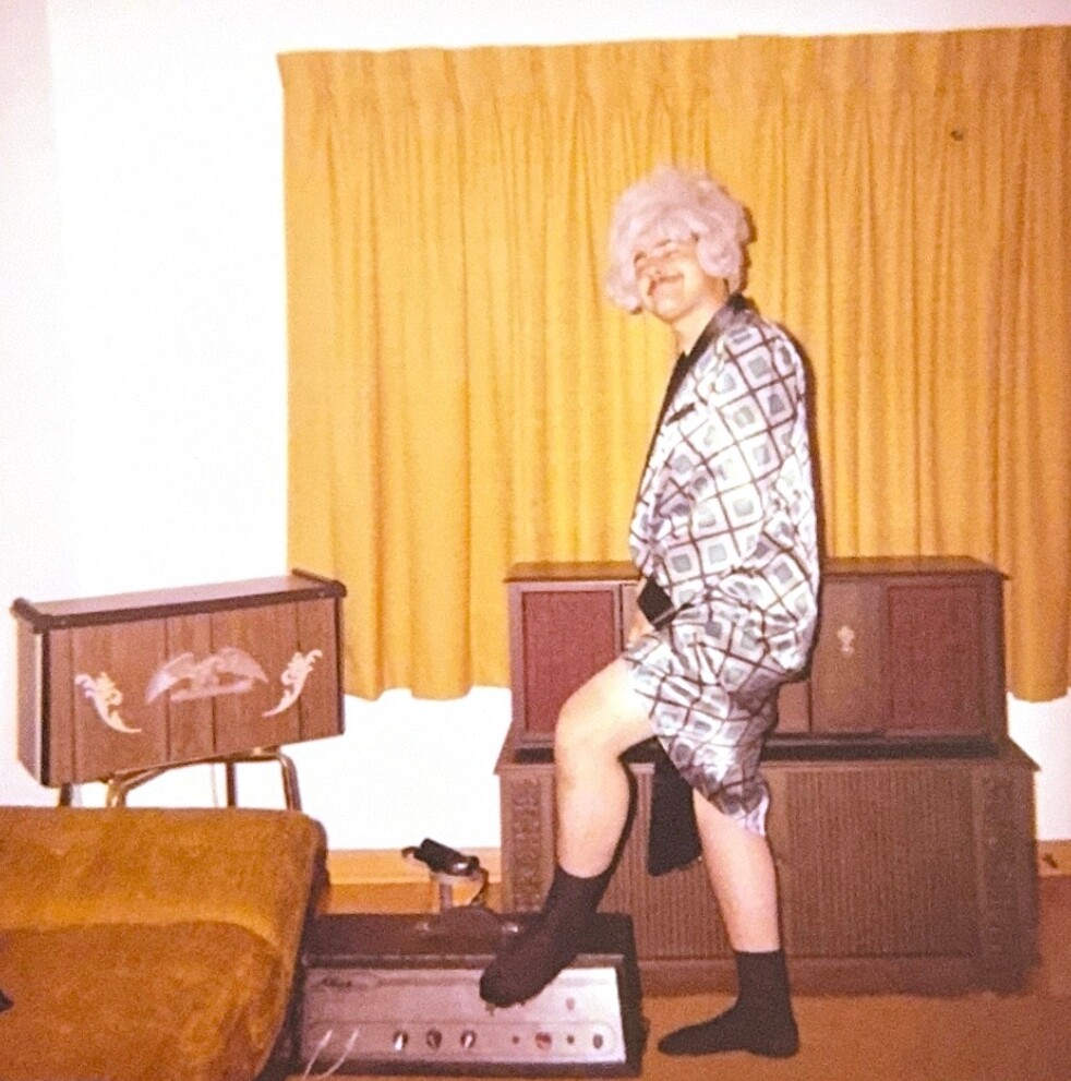 Person in a silver checkered dress and white wig posing with one foot on a vintage amplifier in a room with mustard-yellow curtains.