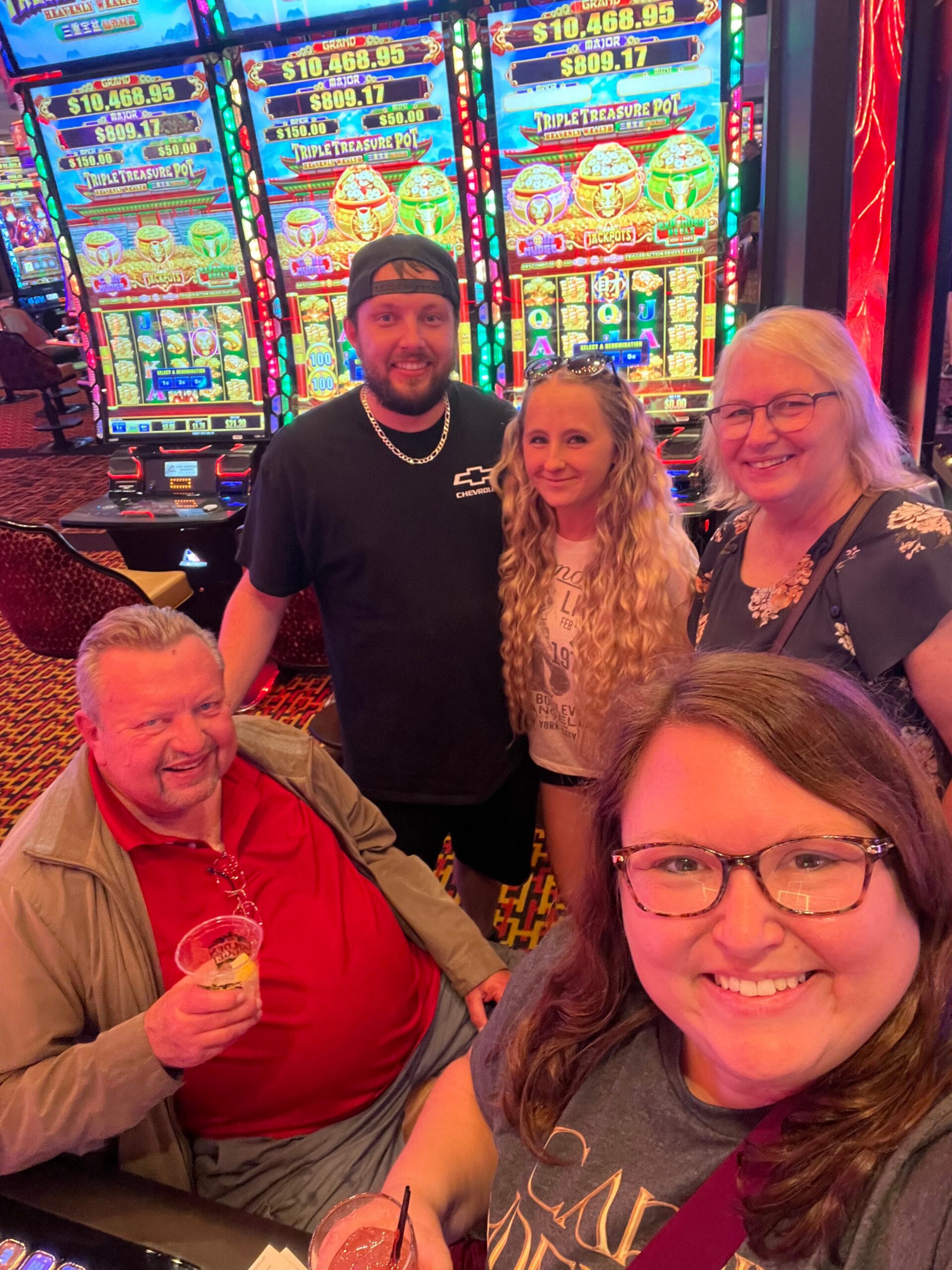 Group of smiling people posing for a selfie at a row of colorful slot machines in a casino, one holding a drink in front of bright screens.