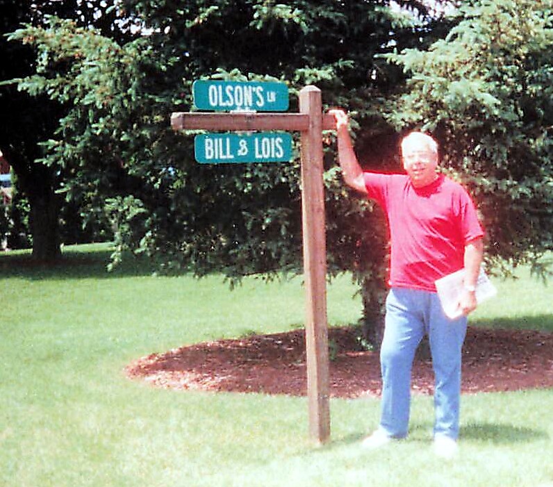 Man in a red shirt standing beside a wooden signpost with two blue street signs reading Olson's Ln and Bill & Lois in a grassy park.