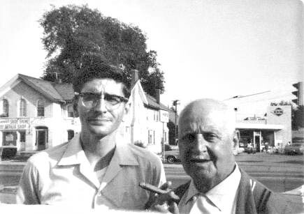 Two men stand on a street corner; one wears glasses and has a cigarette, the other is older and smiling, with storefronts in the background.