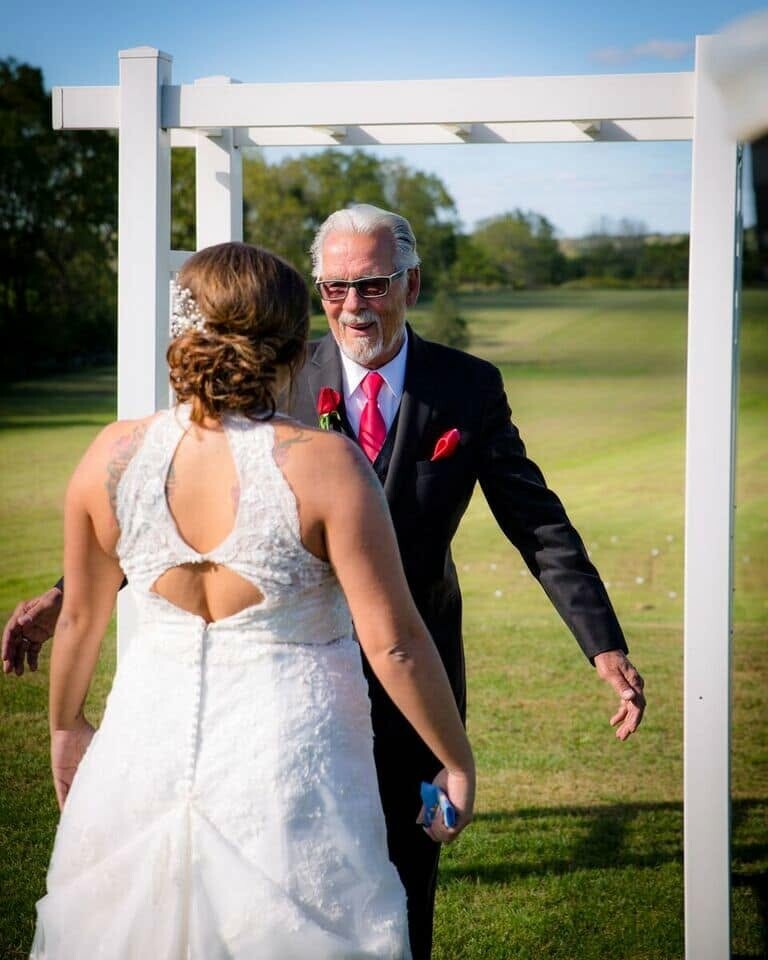 Bride in a white lace wedding dress facing an older man in a dark suit and pink tie outdoors beneath a white wooden arch, about to greet him at a ceremony site.