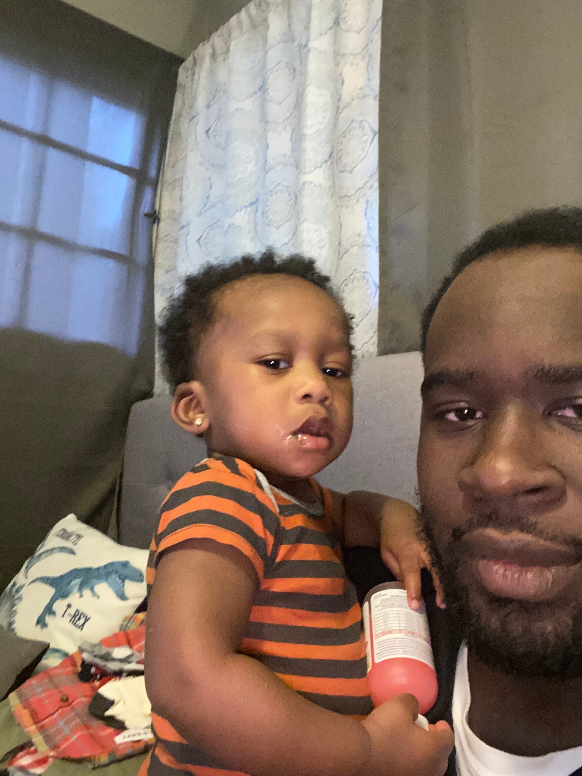 A man with a beard holding a young child wearing an orange and gray striped shirt, indoors with a curtain and window in the background; child holds a pink bottle close to the camera.