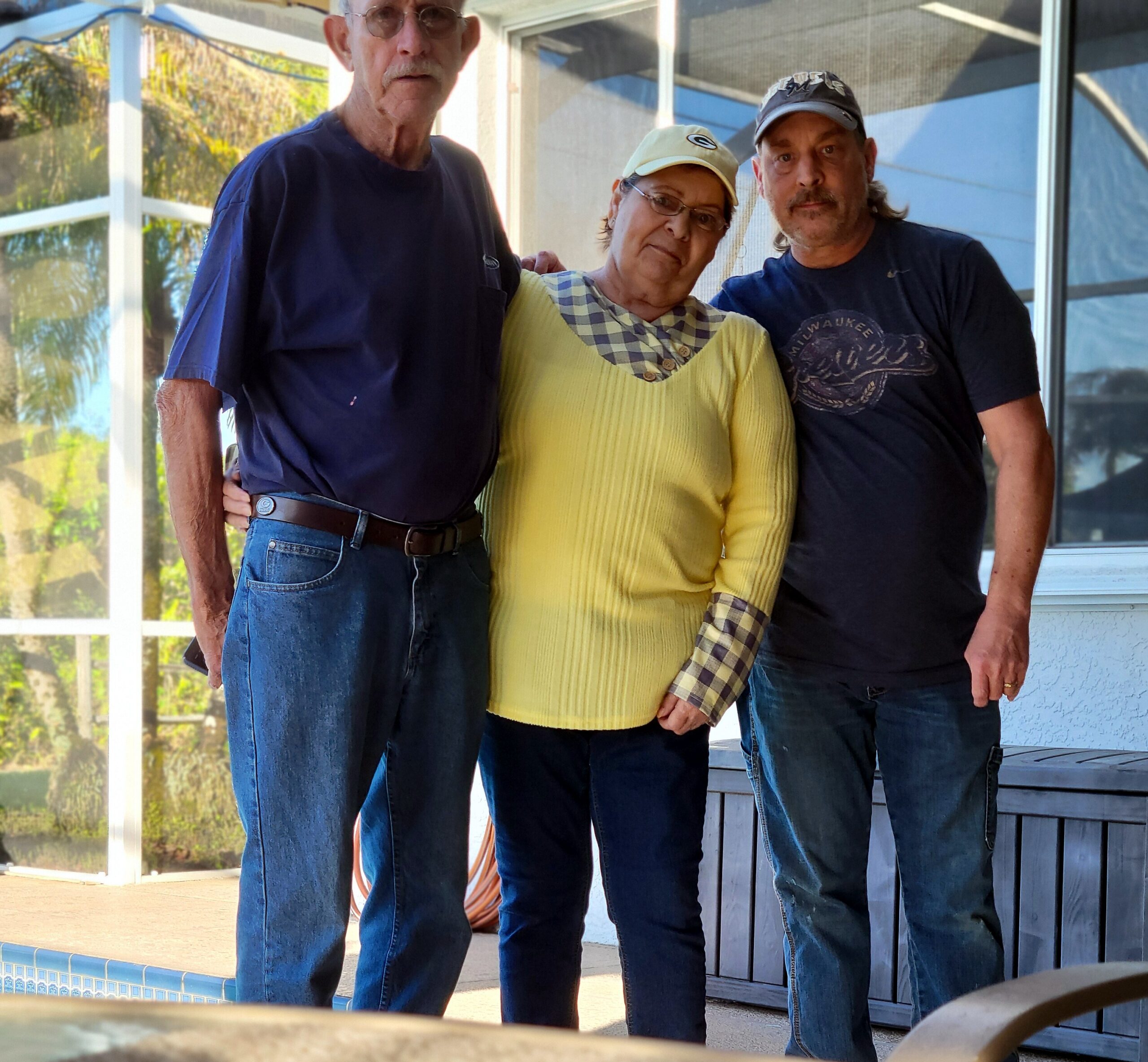 Three adults posing together outdoors by a house and pool: left man in blue shirt, center woman in yellow sweater and cap, right man in dark shirt with a cap.