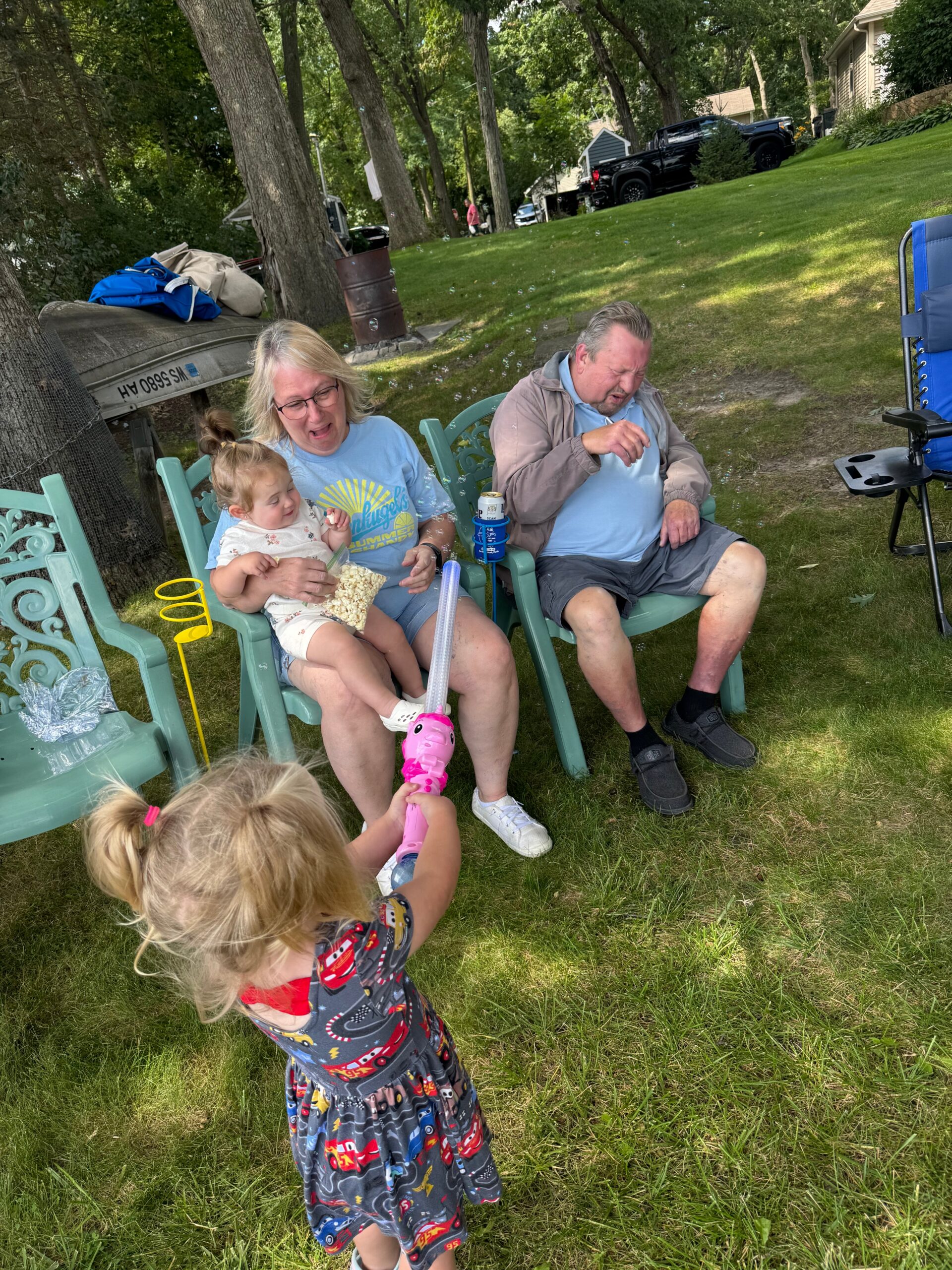 Grandparents sit in green lawn chairs in a shaded park as a toddler runs up with a pink bubble wand and a young girl snack on the woman's lap nearby.