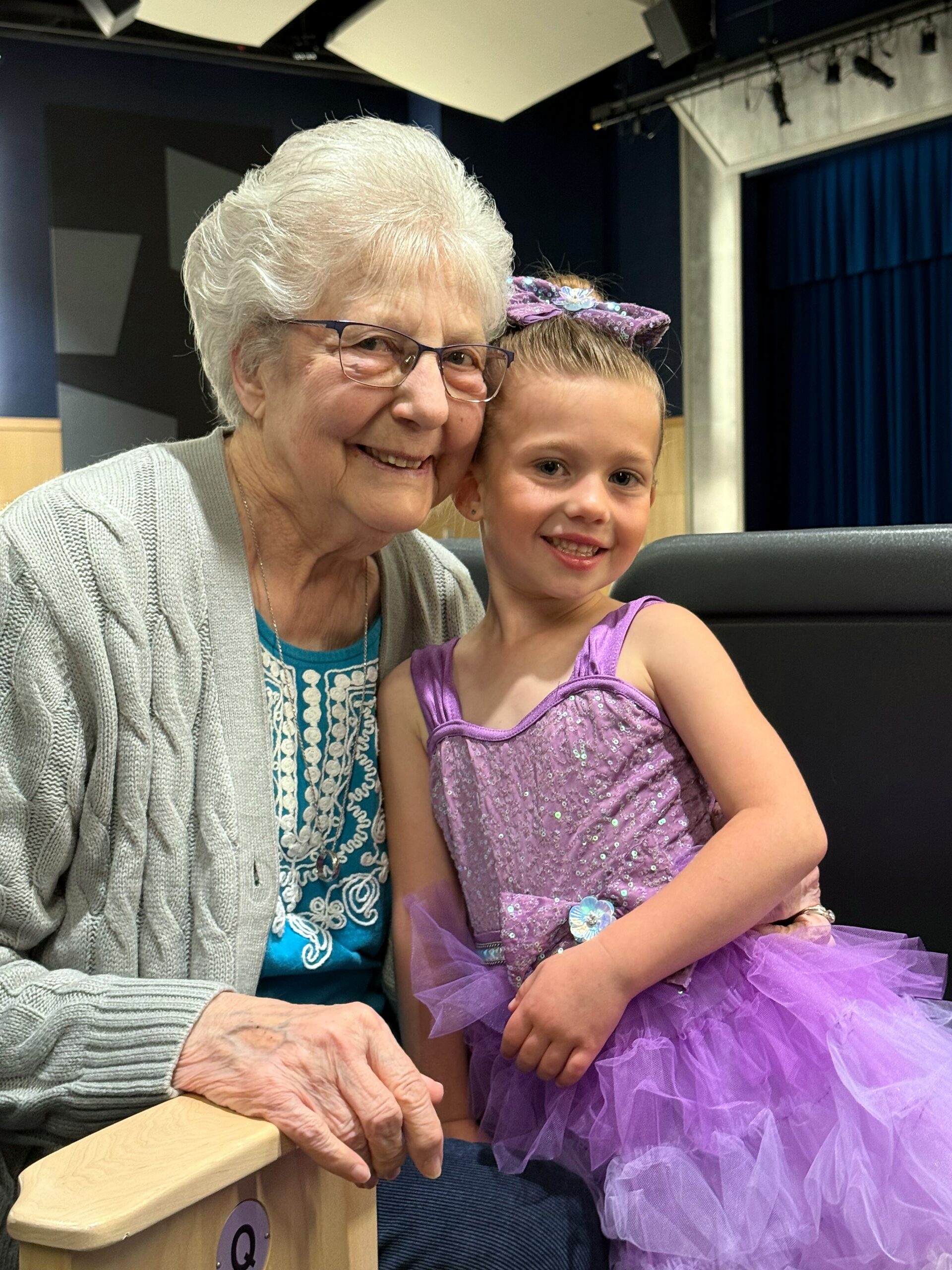 Grandmother and young girl smiling together, seated backstage with stage backdrop behind them.