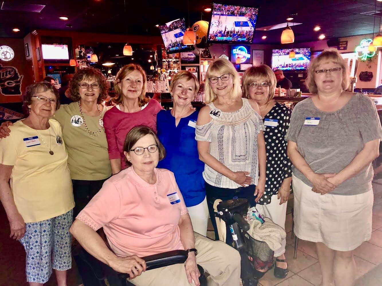 Group of eight women posing together in a casual restaurant, several wearing name badges; one woman sits in a wheelchair in front.