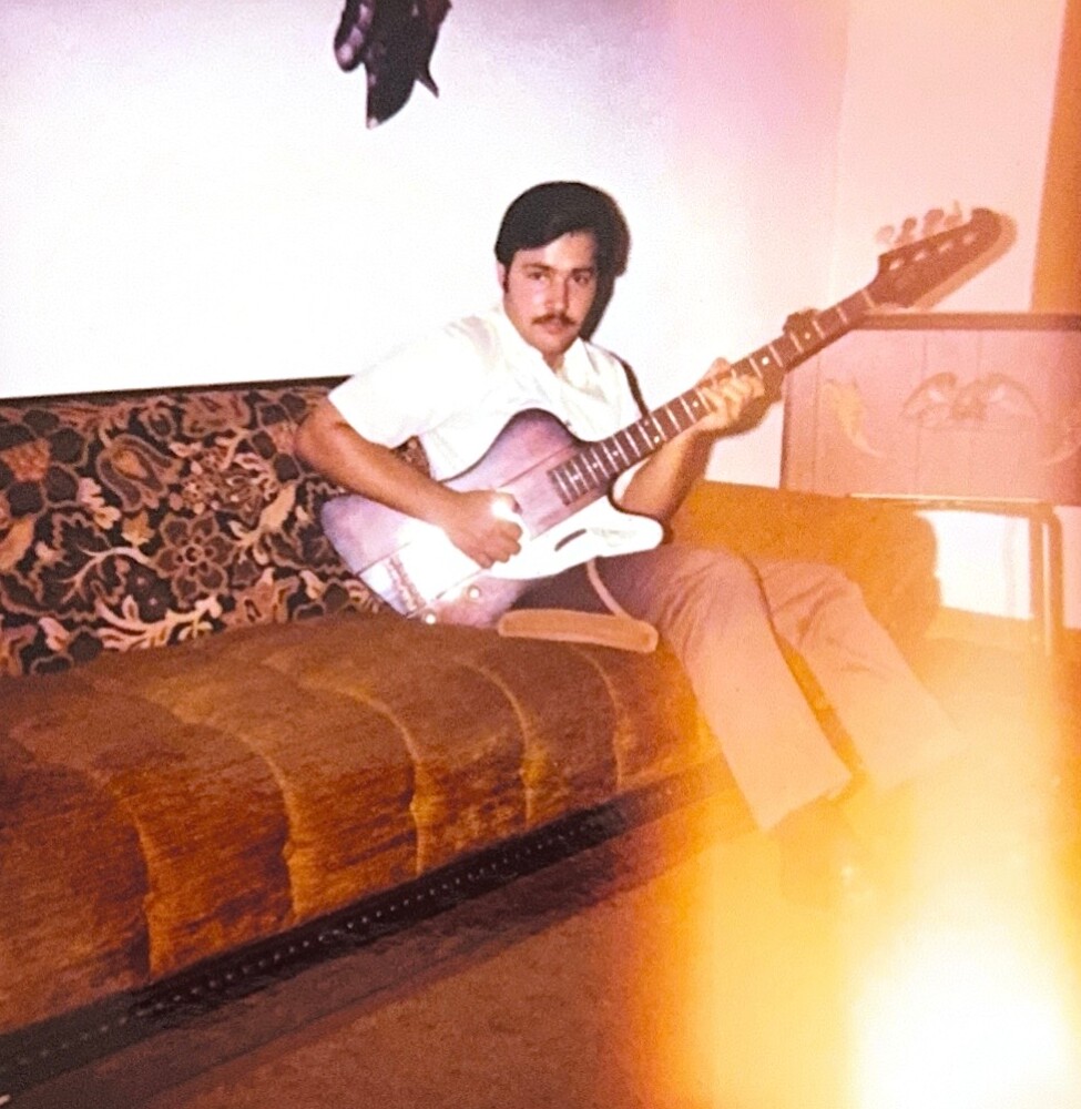Man with dark hair and mustache sits on a patterned sofa, playing a pink electric guitar indoors.