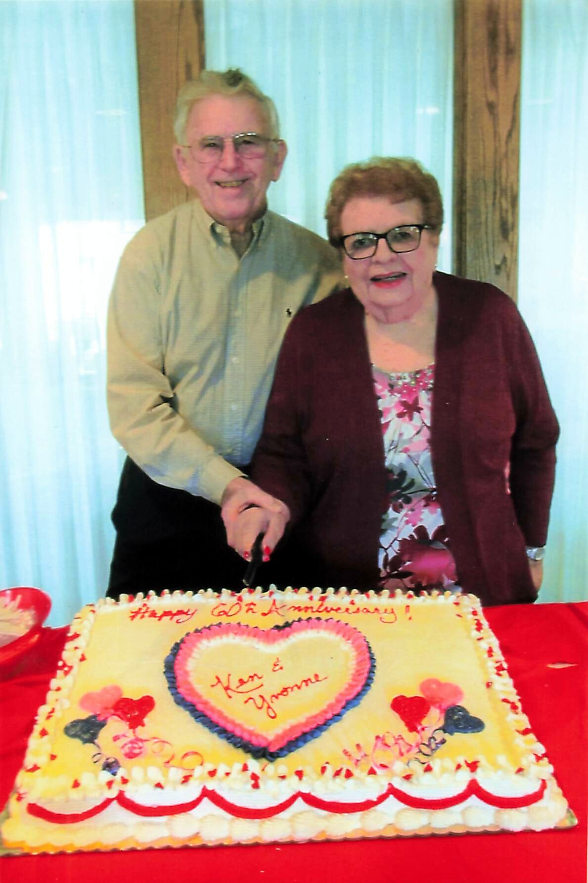 Smiling elderly couple cutting a large rectangular anniversary cake with a heart design at a red-draped table.