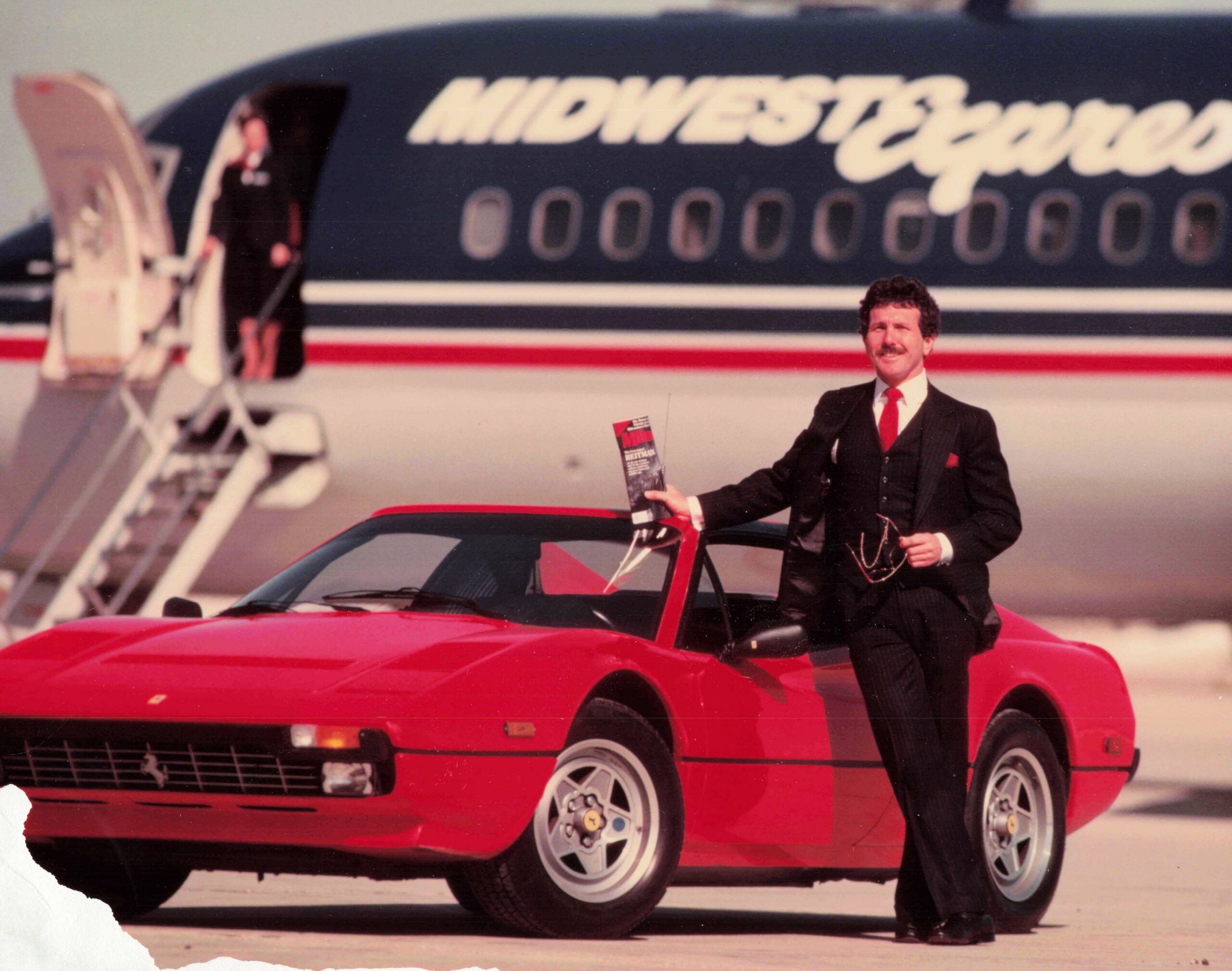 Man in a dark suit with a red tie posing beside a bright red convertible on a tarmac, with a blurred aircraft in the background read 'Midwest Express'.