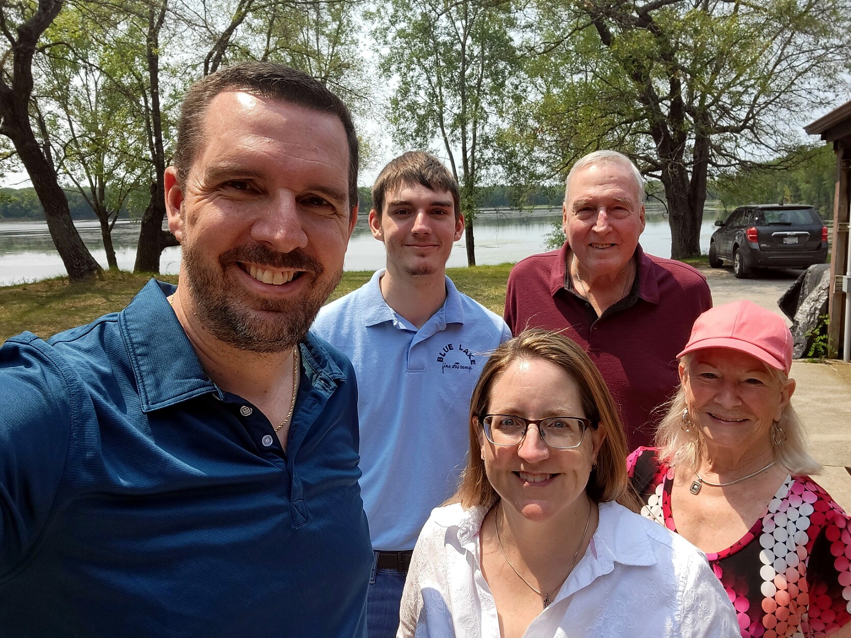 Five adults smiling at the camera outdoors by a lake, with trees and a sunny sky in the background.