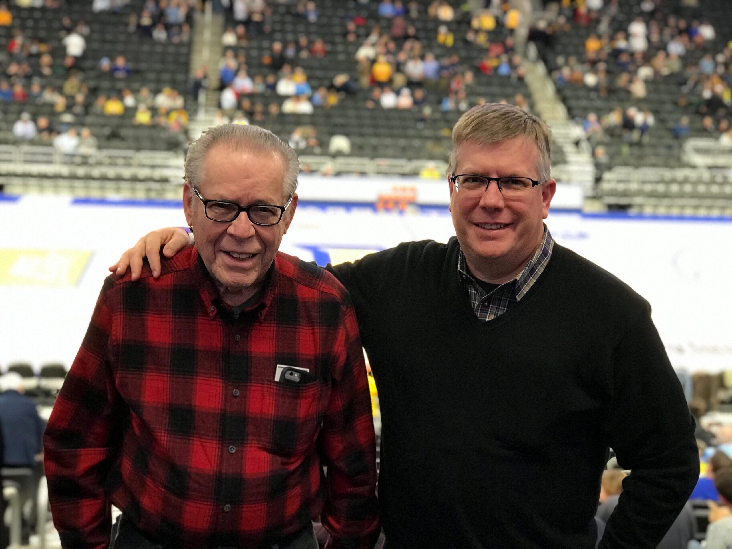 Two men pose with arms around each other at an indoor arena, one in a red plaid shirt and the other in a black sweater, smiling with a crowd in the background.