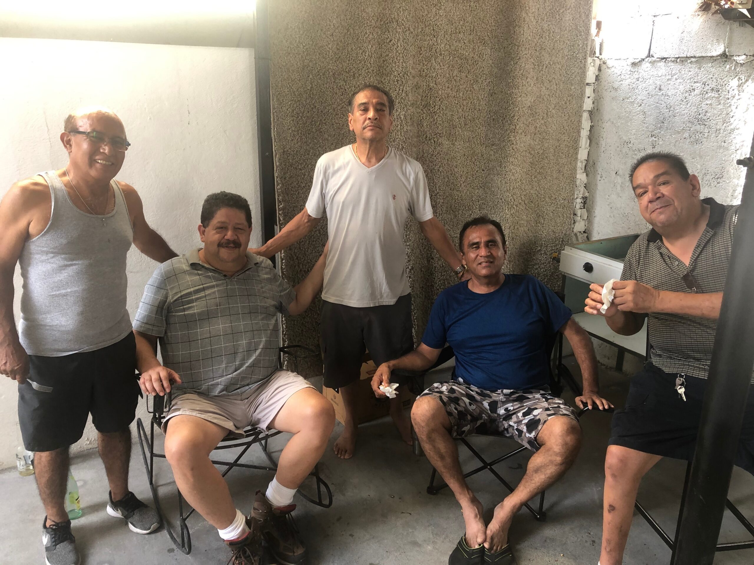 Six men pose together in a concrete-walled room, some seated in foldable chairs, casual clothing and relaxed smiles, suggesting a casual gathering.