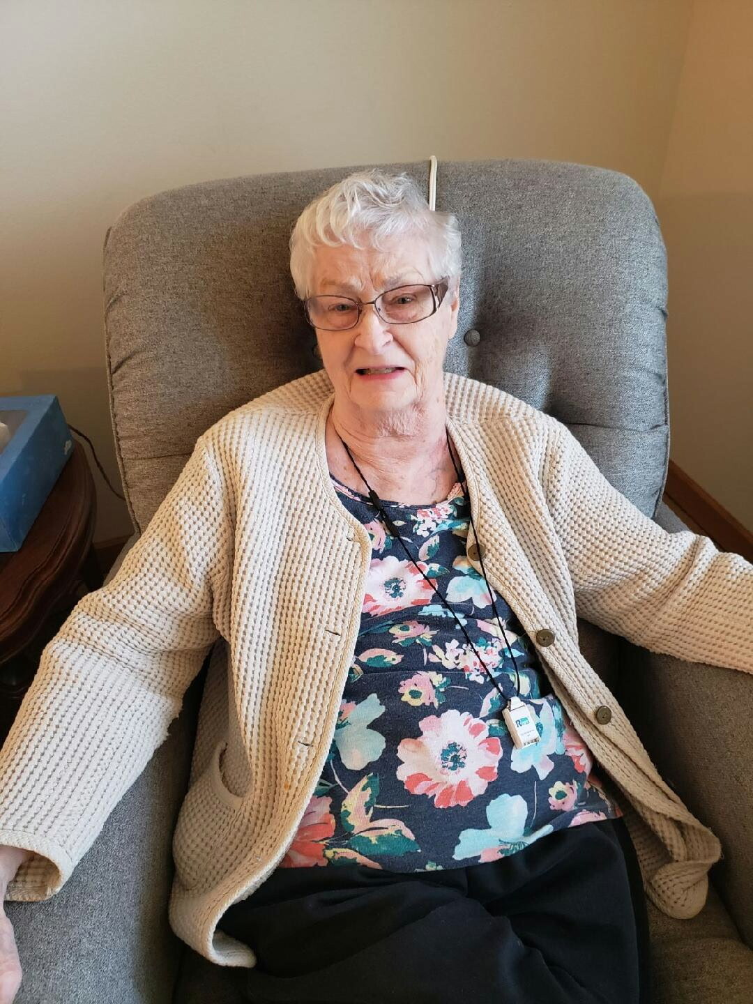 Senior woman with short white hair and glasses sits in a gray armchair, wearing a beige cardigan over a floral blouse.