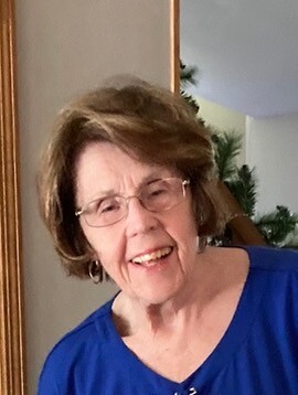 Older woman with short brown hair and glasses, wearing a blue blouse, smiling indoors.