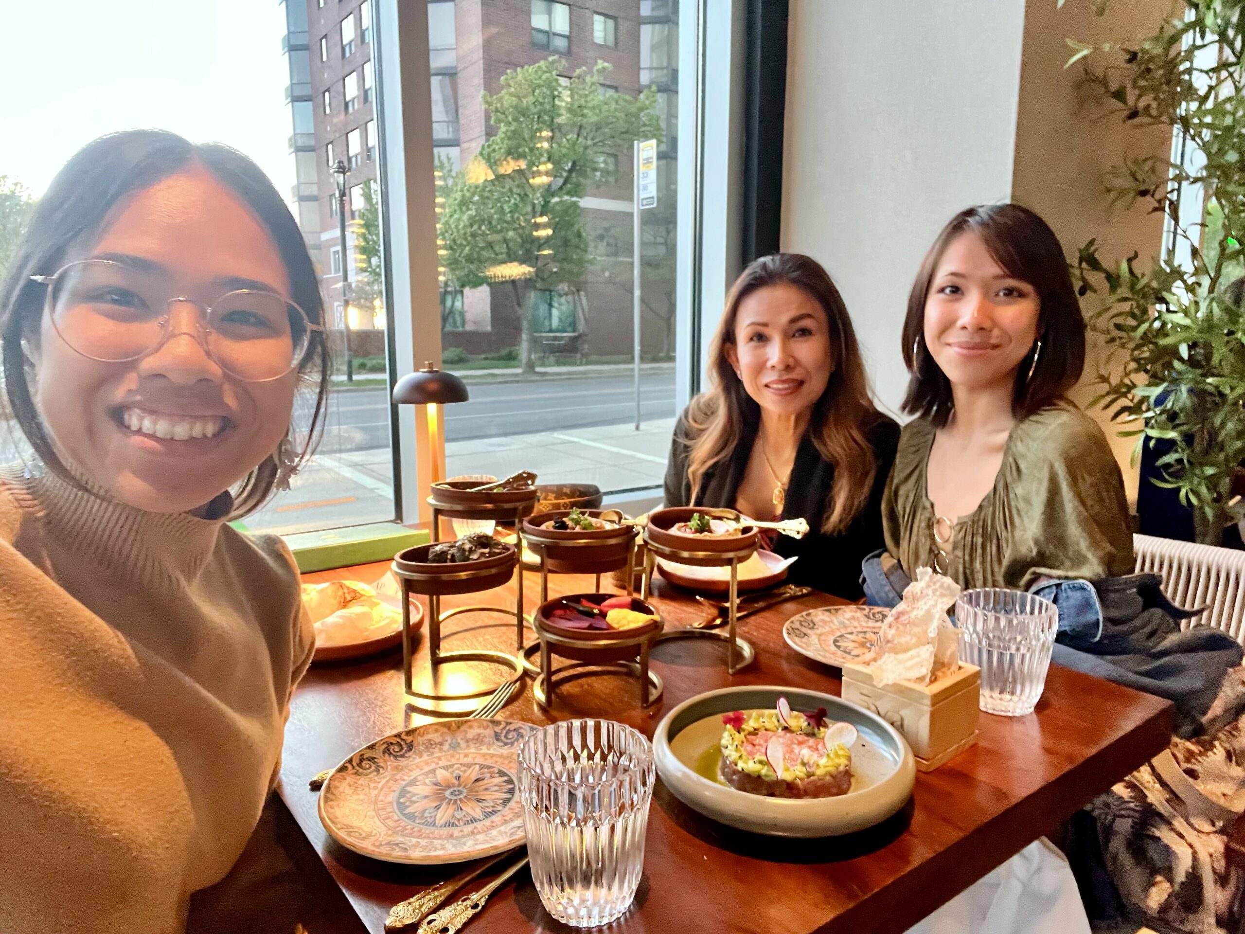Three women at a cafe table posing for a selfie with dessert towers and plates in front of them by a large window.