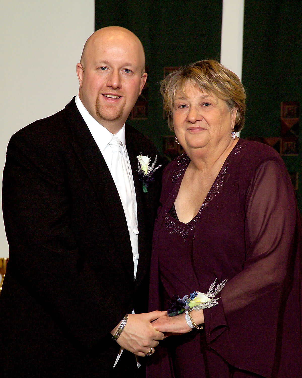 Couple posing for a formal event photo: man in a black tuxedo with white boutonniere beside a woman in a plum dress with a corsage, both smiling at the camera.