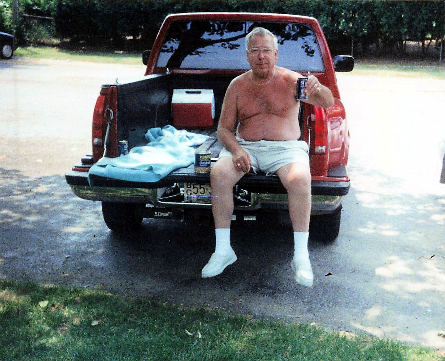 Man in shorts and white socks sits on the tailgate of a red pickup truck, drinking from a can with a towel and cooler in the bed nearby.
