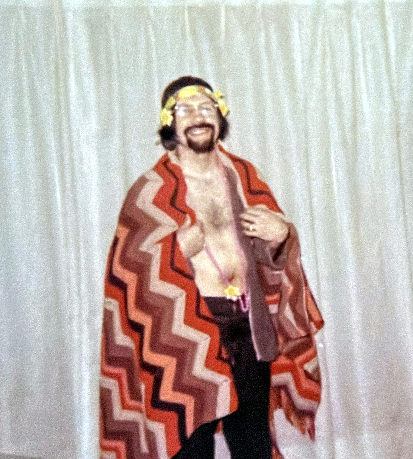Smiling man wearing a multicolored zigzag poncho and flower crown on a stage with a white curtain backdrop.