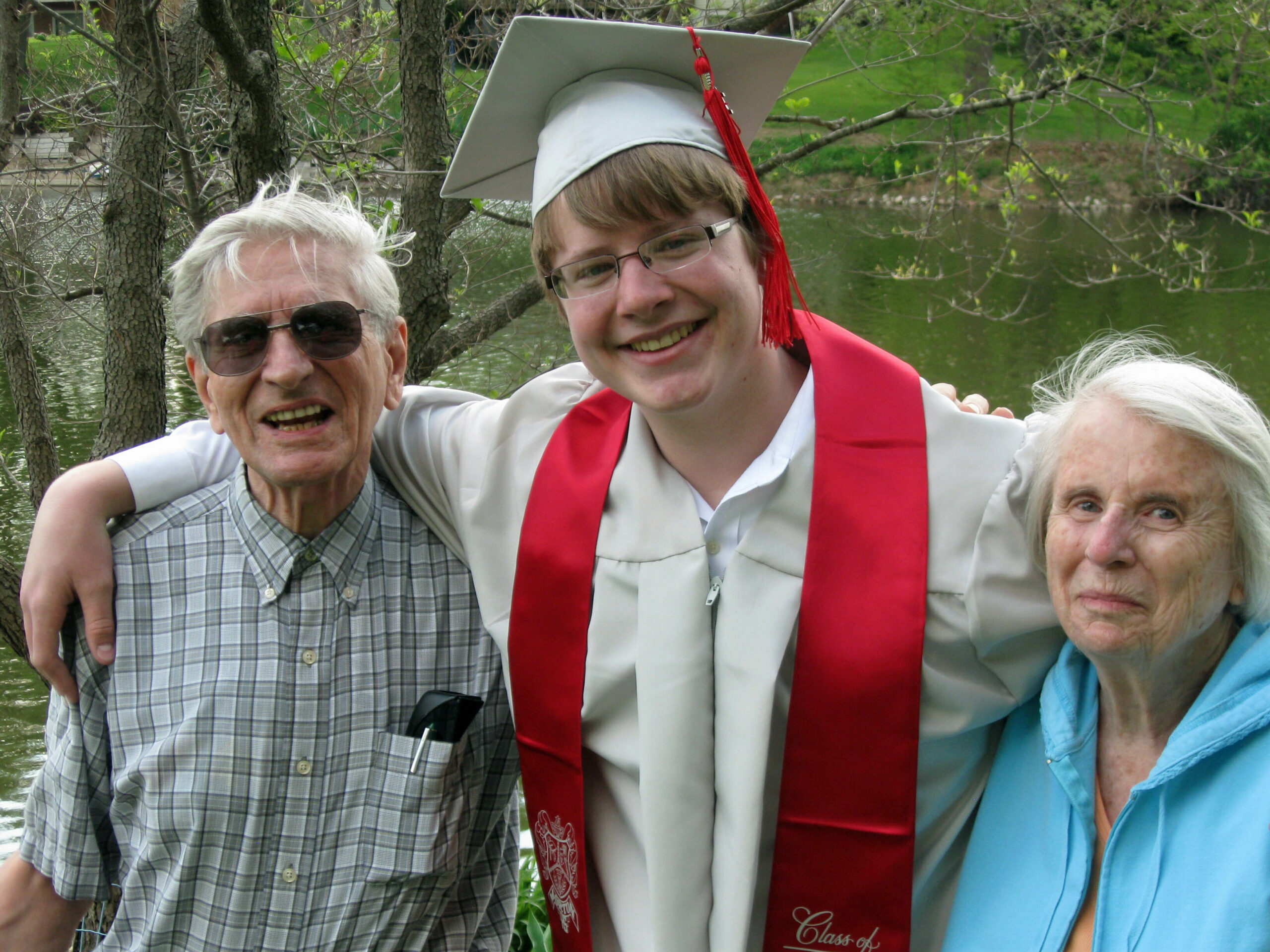 Graduate in a gown and cap with a red stole posing with two seniors outdoors by a pond or lake.
