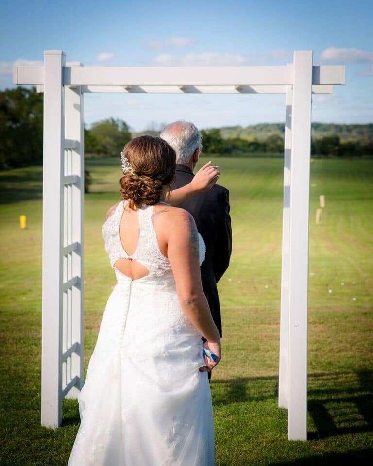 Bride in a white wedding gown stands with an older man under a white garden arch on a sunny grassy field.