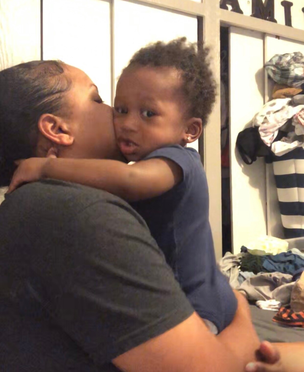 Adult caregiver holding and kissing a young child on the cheek in a cluttered bedroom.