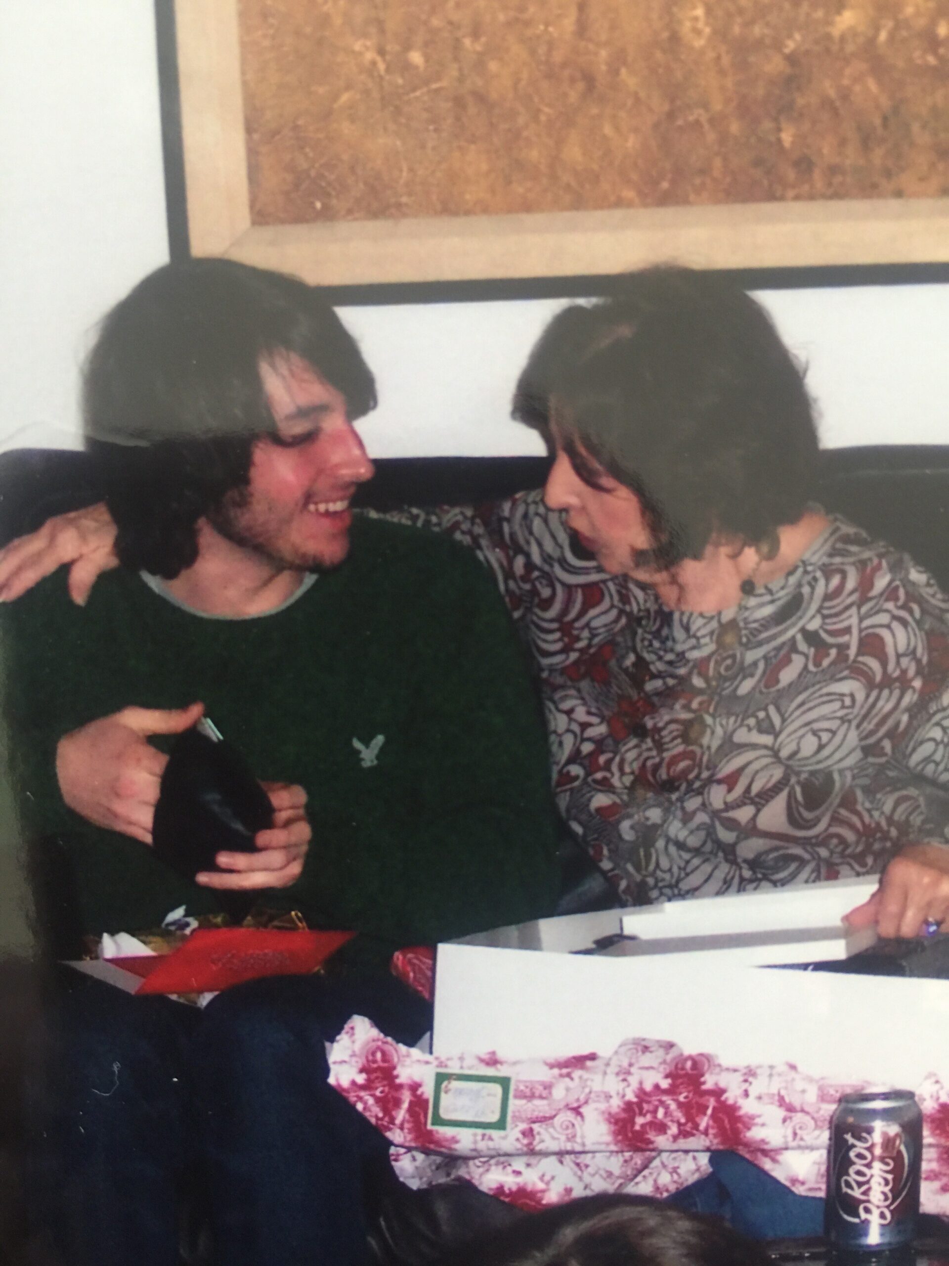 Two young men sit close on a couch, smiling and with arms around each other, enjoying a moment together at a casual gathering.