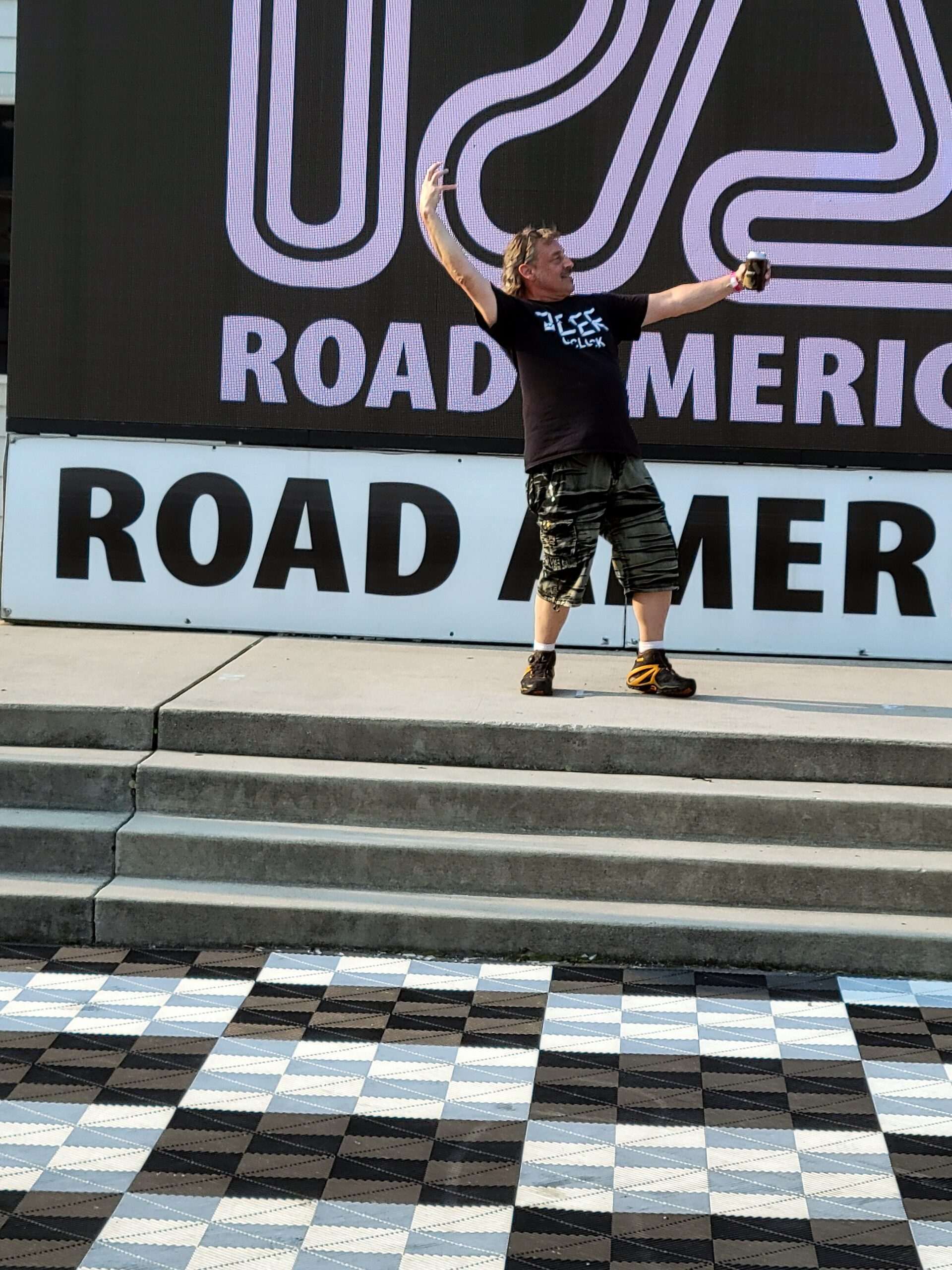 Man in a black shirt and camo shorts stands on steps with arms raised, holding a drink, in front of a large sign behind him.