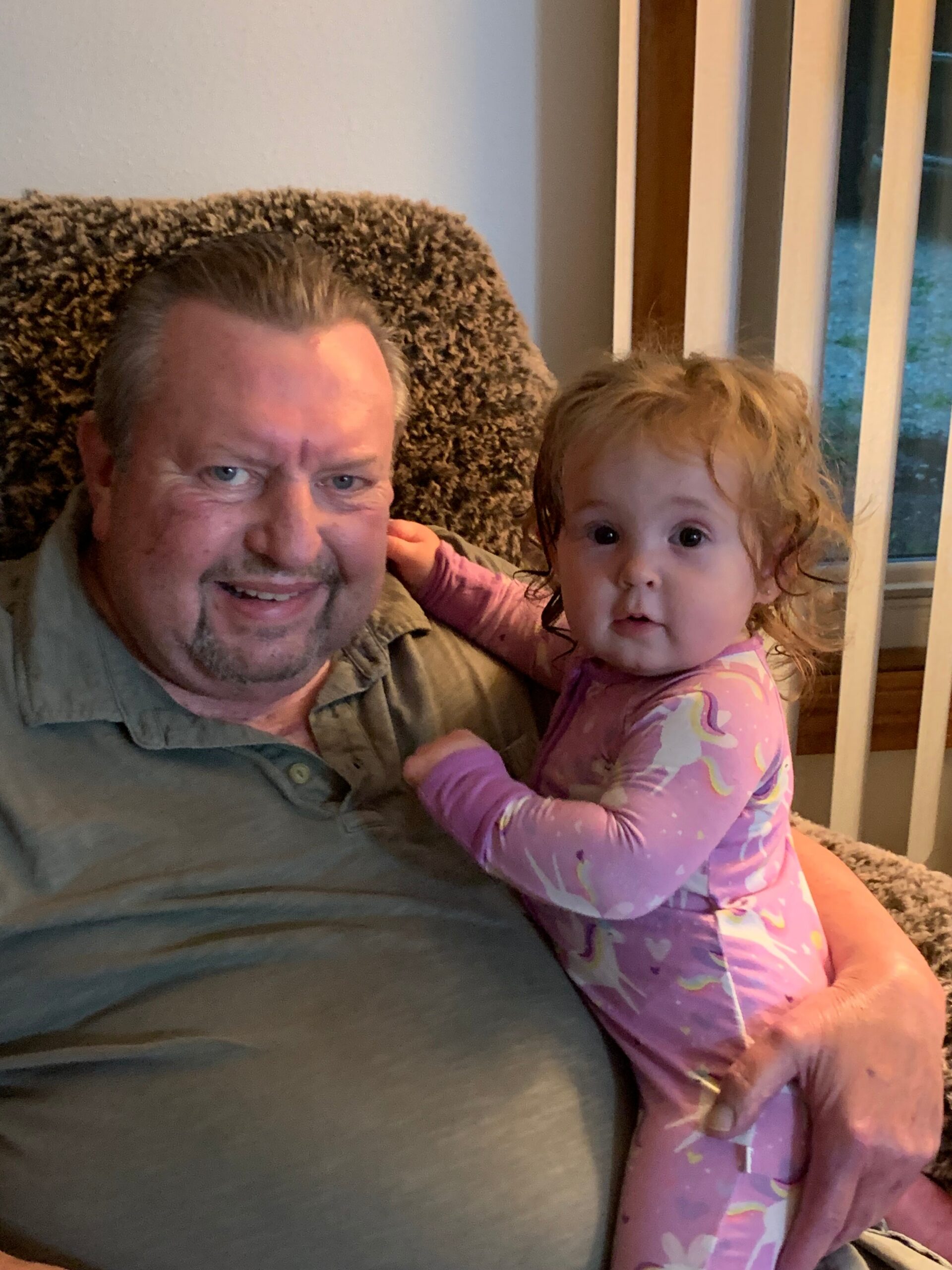 Man and young girl sit closely together on a couch, smiling at the camera, pink pajama-clad child leaning on his chest.