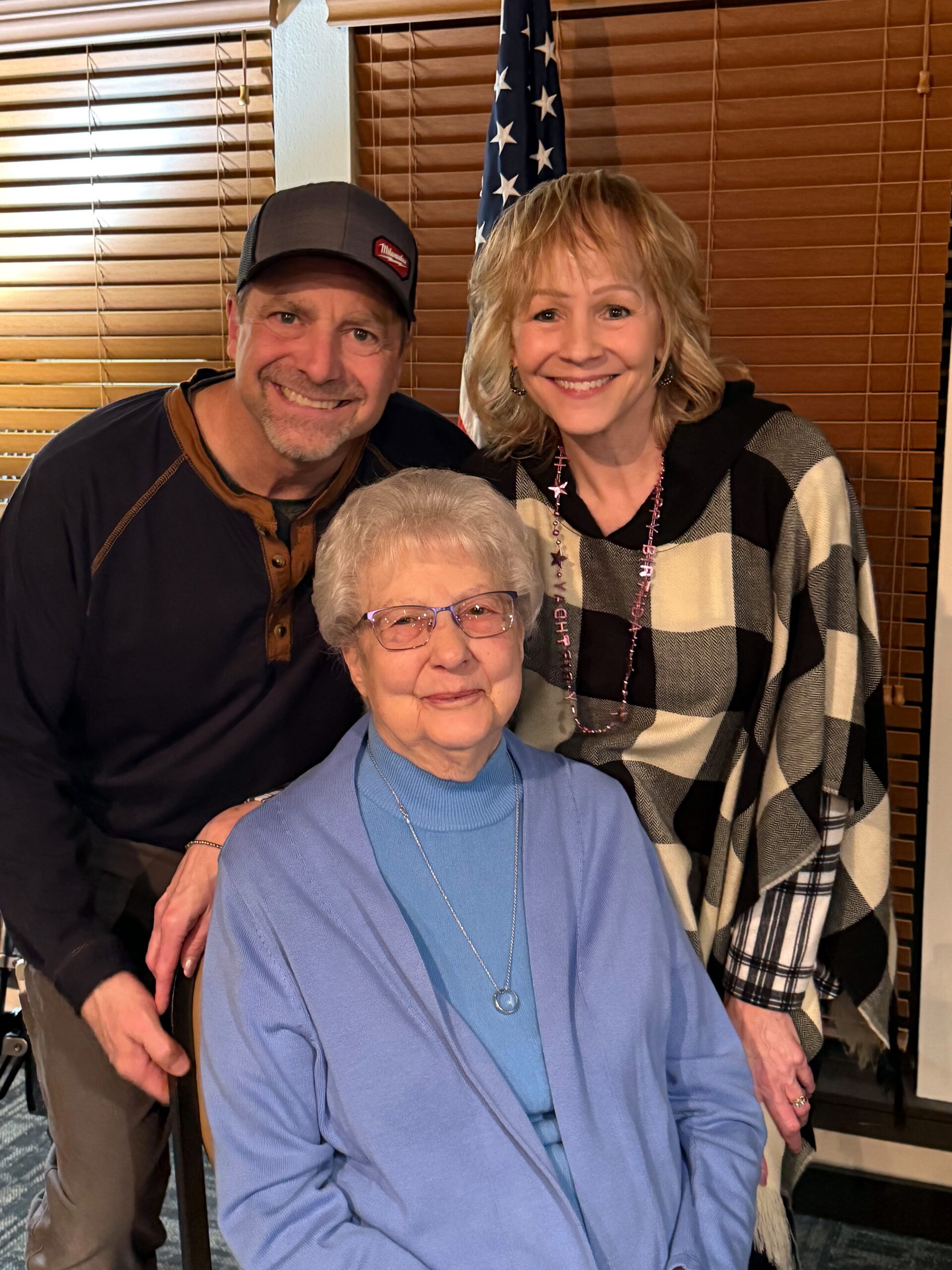 Three people smile for a family portrait indoors; an American flag is in the background.