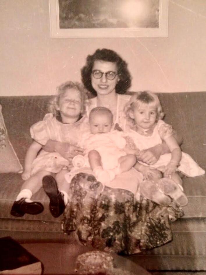 Vintage family portrait: a woman with glasses sitting on a couch, flanked by two young girls holding a baby between them, all smiling.