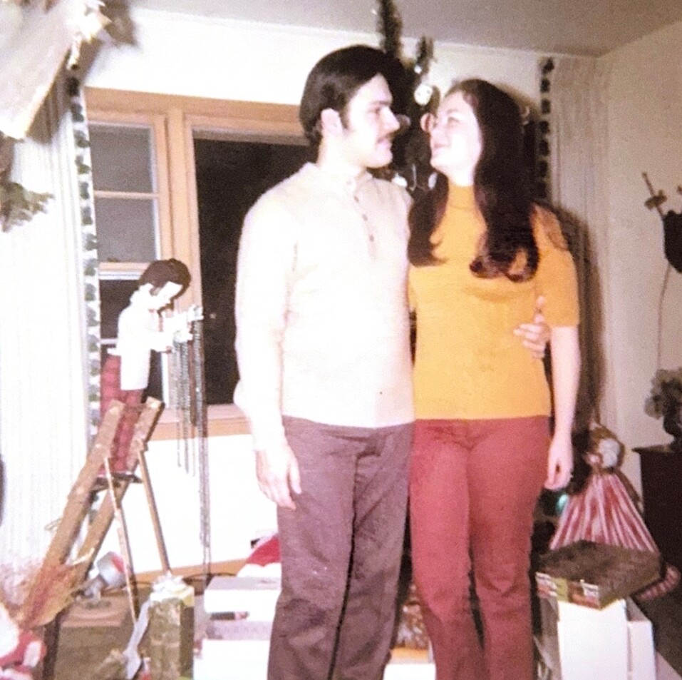 A couple stands close together and smiles at each other in a festively decorated living room during Christmas season.