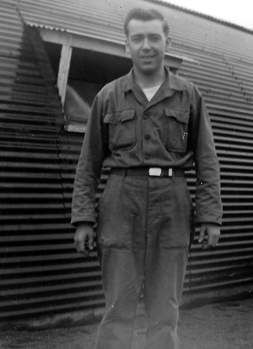 Man in a utility jumpsuit stands outdoors in front of a corrugated metal wall.