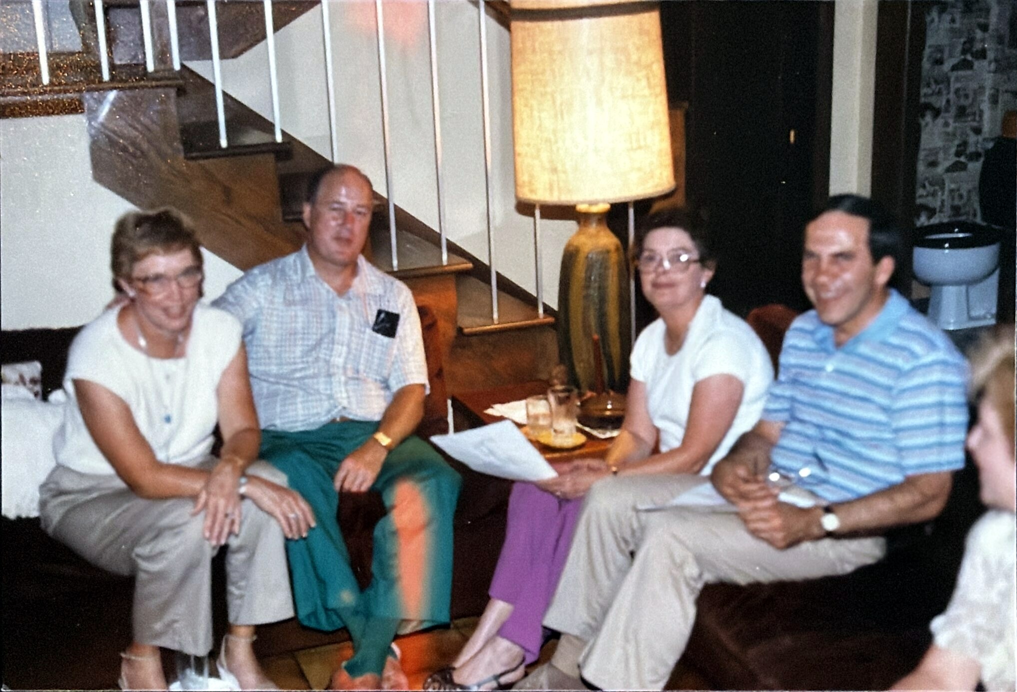 Four adults sit together on a sofa in a cozy living room, smiling at the camera with drinks and papers on the table in front of them.