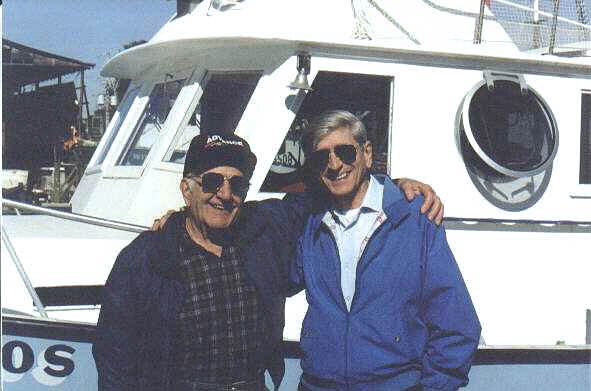 Two men stand close together in front of a white boat, smiling with arms around each other on a dock.