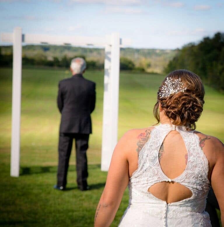 Bride in a white lace dress with an updo watches her groom at an outdoor wedding beneath a white arch on a grassy field.