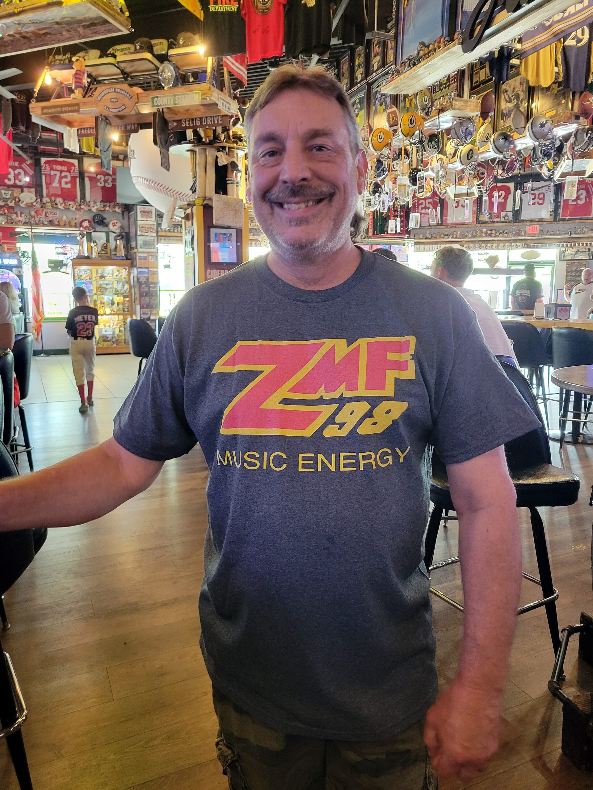 Smiling man wearing a gray 'ZMF 98 Music Energy' shirt in a sports bar filled with jerseys and memorabilia.