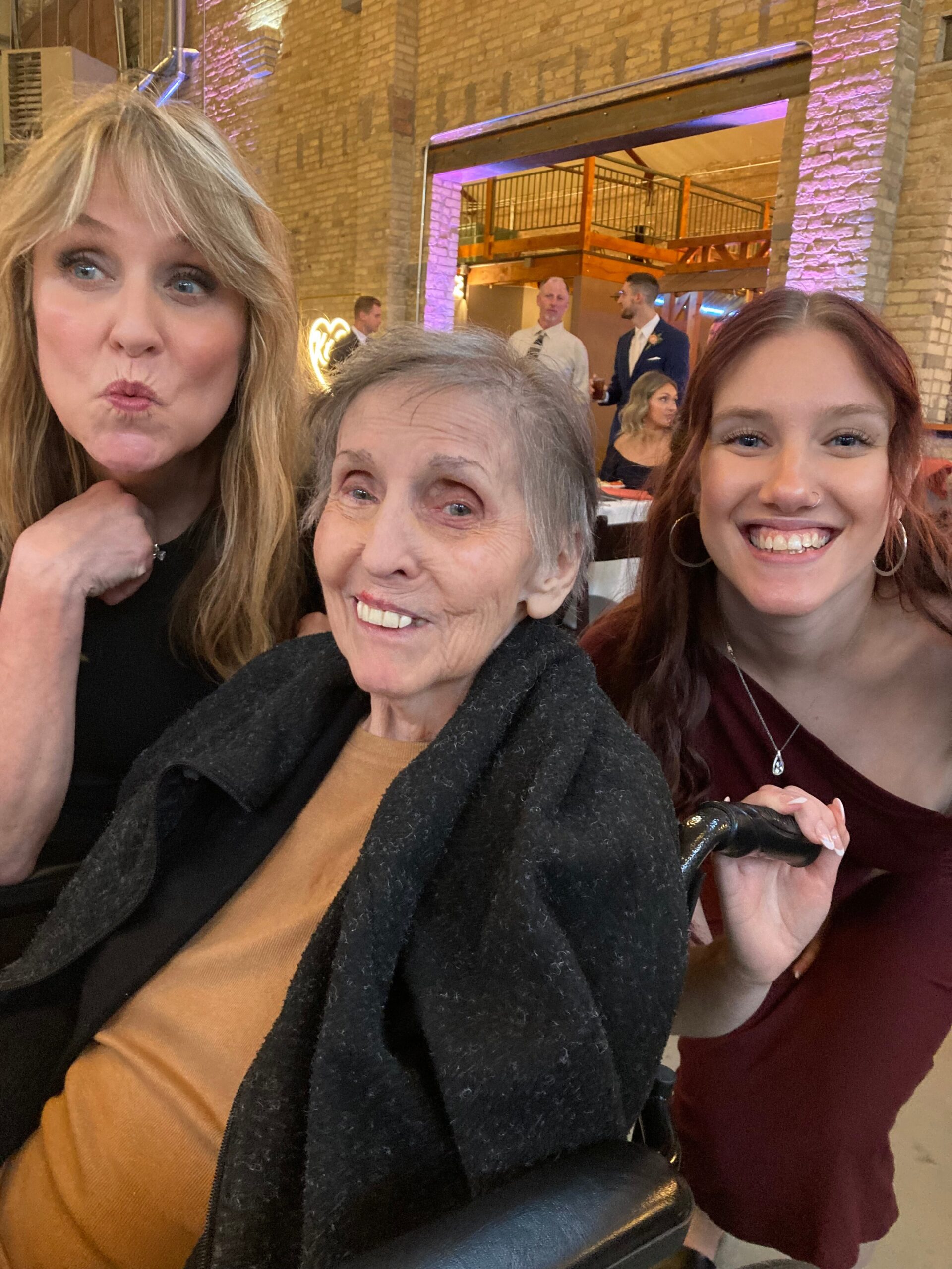 Three women pose for a close selfie at an indoor event—left woman puckers her lips, center elderly woman smiles, right smiling young woman with red hair in a burgundy dress. Background shows brick walls and warm purple lighting.