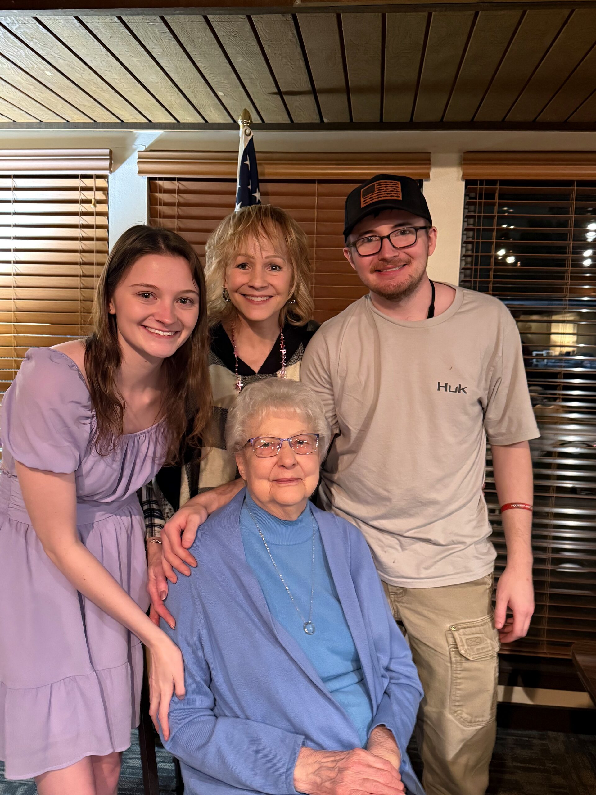 Family portrait indoors: elderly woman in blue cardigan seated center, flanked by a smiling young woman in lavender dress and a young man in a cap, an American flag behind them.