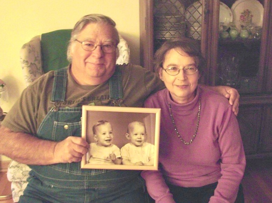 Elderly man in overalls and a woman in a purple sweater sit together on a couch, holding a framed photo of two smiling babies.