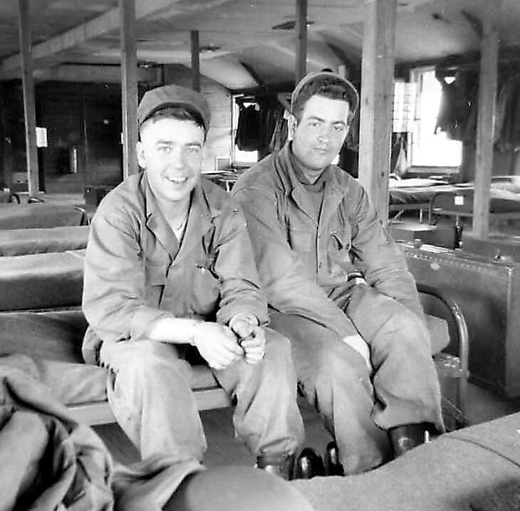Two soldiers in uniform sit on a bench inside a wooden barracks, smiling at the camera.