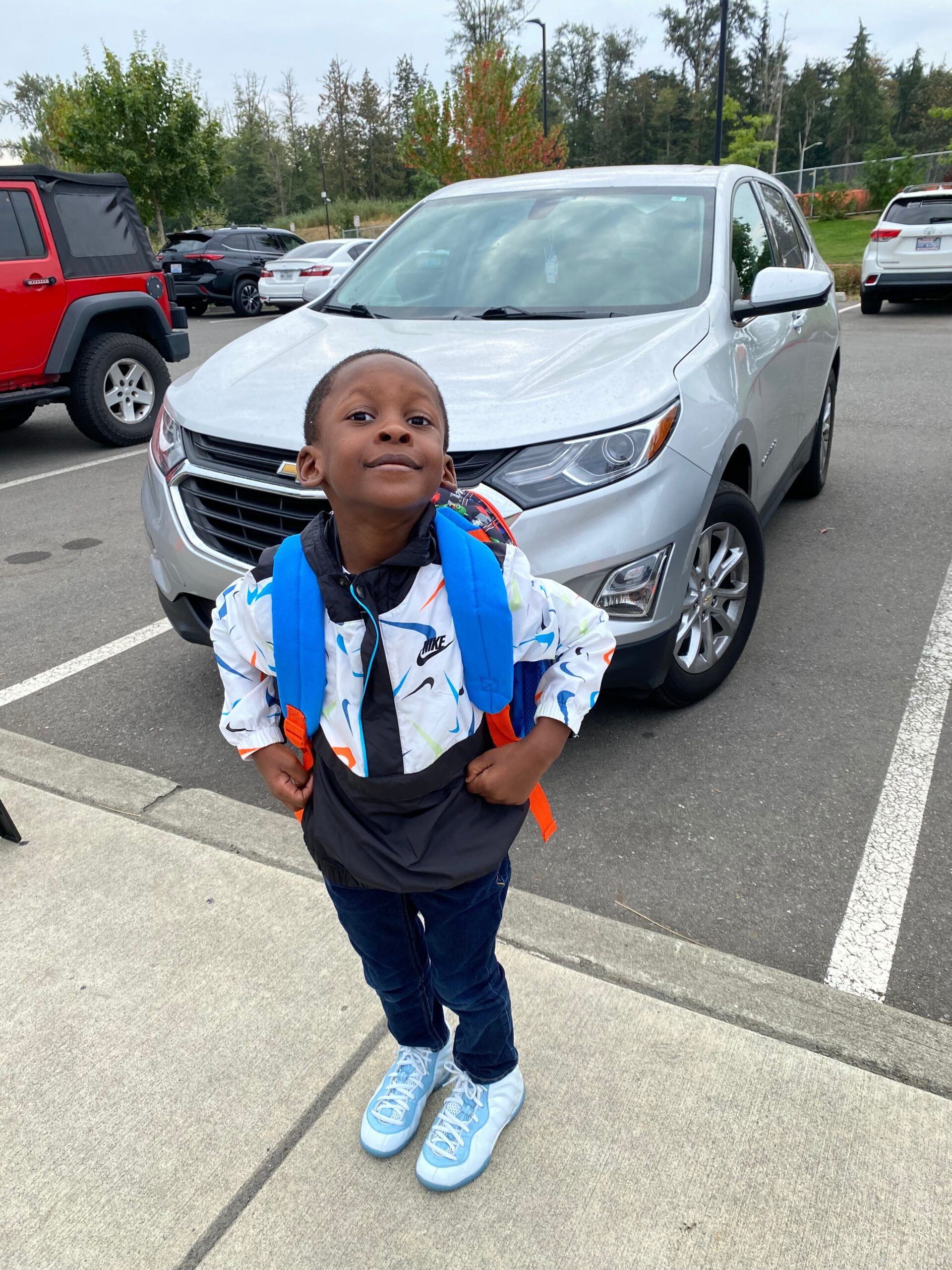 Smiling boy with a blue backpack posing in a parking lot beside a silver SUV/car.