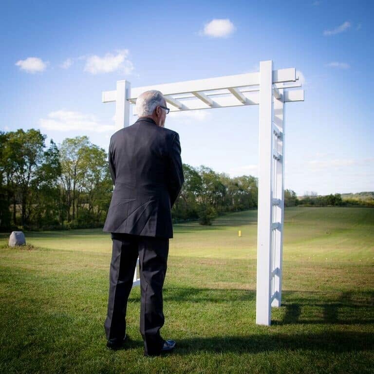 Older man in a black suit stands with his back to the camera, gazing at a white wooden arch on a sunny grassy field.