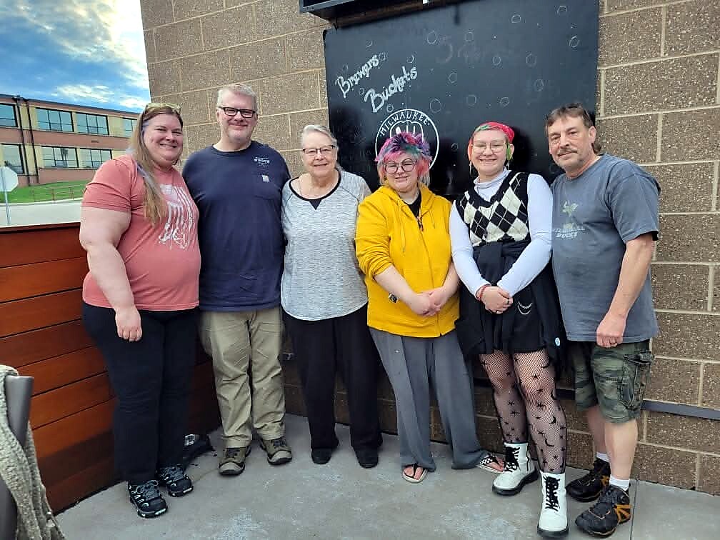 Six adults standing in a line outside a building, posing for a group photo near a chalkboard sign.